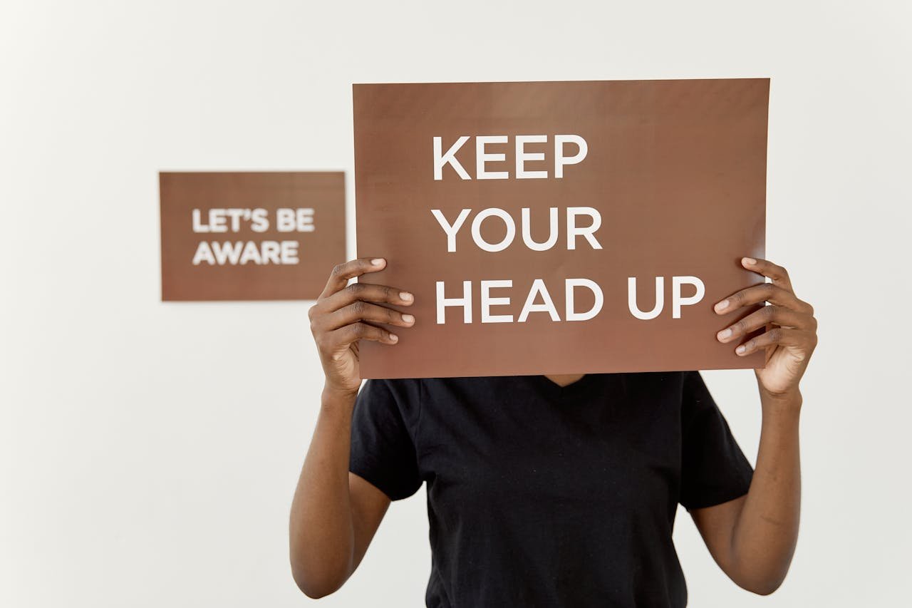 A motivational sign reading KEEP YOUR HEAD UP held by a person indoors.